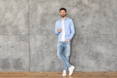 Mavi düğmeli gömlekli ve kot pantolonlu bir adam bir bacağı diğerinin önünde çapraz duruyor, gri beton bir duvara yaslanıyor. Elinde bir akıllı telefon tutuyor.