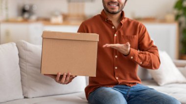 Kırpılmış bir adam oturma odasındaki beyaz bir kanepede oturuyor, önünde sol eliyle bir karton kutu tutuyor. Gülümsüyor ve sağ eliyle hareket ediyor.