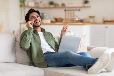 Yeşil düğmeli gömlek ve kot pantolon giyen genç bir Arap bir kanepede oturmuş dizüstü bilgisayar tutarken telefonla konuşuyor. Gülümsüyor ve yukarı bakıyor. Rahatlamış ve zamanının tadını çıkarıyor..