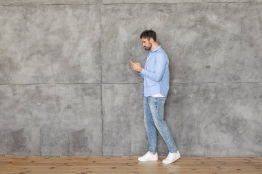 Açık mavi gömlekli ve kot pantolonlu bir adam akıllı telefonuna bakarken beton bir duvarın yanından geçiyor. O, beyaz spor ayakkabı giyiyor. Zemin ahşap, kopyalama alanı