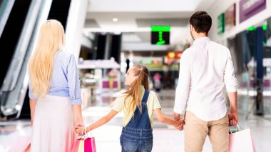 Üç kişilik bir aile alışveriş merkezinden geçiyor. El ele tutuşuyorlar ve ebeveynlerin her biri bir alışveriş çantası taşıyor. Genç kız arkalarından bir şeye bakıyor.