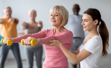 Genç bayan koç, yaşlı kadına yardım ederken halterlerle egzersiz yapıyor. Yaşlılar için spor salonundaki spor dersine gidiyor, eğitmenle antrenman yapıyor.
