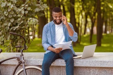 Parktaki belgelere bakarken bankta oturan ve cep telefonuyla konuşan yakışıklı afro adam, ofis dışında çalışıyor.