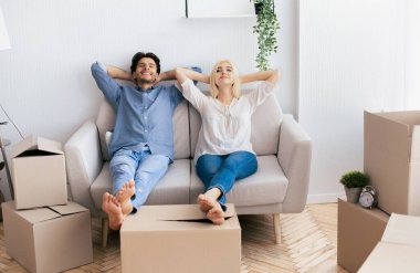 Rahat bir çift, yeni evlerindeki kanepeye oturur. Etrafı karton kutularla çevrilidir..
