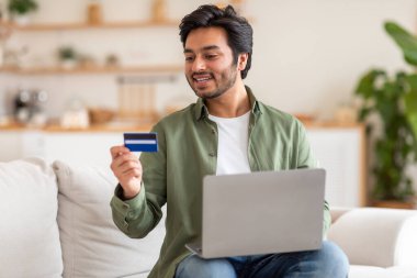 Genç bir Arap adam evindeki beyaz bir kanepede oturuyor, sağ elindeki dizüstü bilgisayara bakarken sol elinde bir kredi kartı tutuyor. Gülümsüyor, tahminen online alımından memnun..