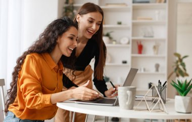 Two Happy Ladies Using Laptop Computer Working And Learning Online Together Or Making Video Call Sitting At Desk In Modern Office. E-Learning. Females Entrepreneurship Career Concept. Side View
