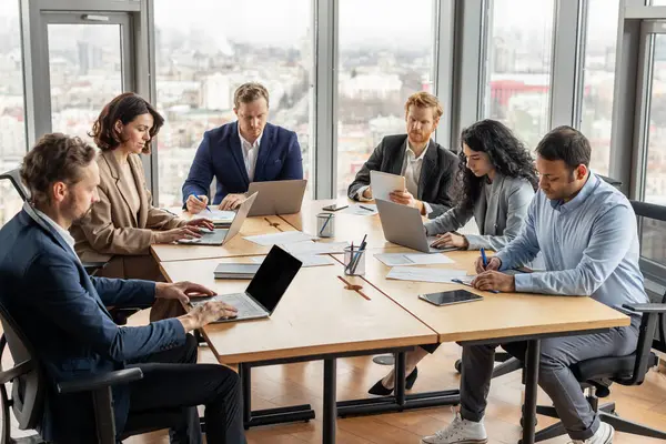 Modern ofis ortamında beş profesyonelden oluşan farklı bir ekip dizüstü bilgisayarlarıyla birlikte çalışıyor. Arkalarındaki büyük pencereler doğal ışık ve şehrin ufuk çizgisini gösteriyor..