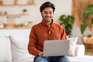 Parlak turuncu tişörtlü Arap bir adam oturma odasında beyaz bir koltukta oturuyor ve dizüstü bilgisayar kullanırken gülümsüyor..