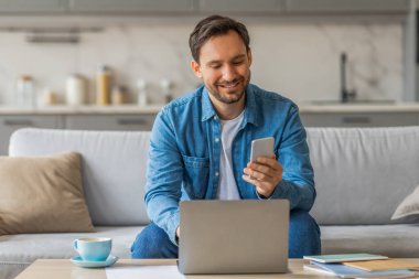 Oturma odasındaki bir kanepede oturan bir adam dizüstü bilgisayarıyla çalışırken aynı zamanda akıllı telefonundan bir şeyi kontrol ediyor. O mavi bir gömlek ve kot pantolon giyiyor. Gülümsüyor, mutlu olduğunu gösteriyor.