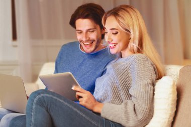 Modern yaşam tarzı. Diz üstü bilgisayar ve dijital tablet kullanan mutlu çift içeride koltukta rahatlıyor. Seçici Odaklanma