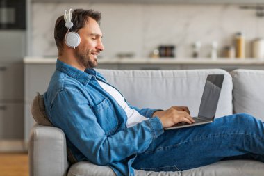 Kot gömlekli bir adam gri bir kanepede bacaklarını açarak oturur. Beyaz kulaklık takıyor ve yazarken dizüstü bilgisayarına bakıyor..