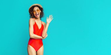 Kıvırcık kahverengi saçlı genç siyah bir kadın kırmızı bir mayo ve güneş gözlüğü takıyor. Gülümser ve kameraya el sallar. Parlak bir turkuazın önünde durur ve uzayı taklit eder.