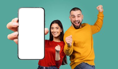 Genç bir çift başarılarını büyük gülümsemeler ve heyecanlı jestlerle kutluyor. Kadın elinde boş bir ekranla akıllı bir telefon tutarken, adam yumruğunu havaya kaldırıyor..