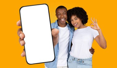 Siyah adam ve kadın sarı arka planın önünde duruyor. Adam elinde boş ekranlı bir akıllı telefon tutuyor. Kadın eliyle 