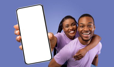 Mutlu bir siyah çift, elinde boş bir ekranla akıllı telefonu tutarken fotoğraf için poz veriyor. İkisi de gülümsüyor ve kameraya bakıyor..