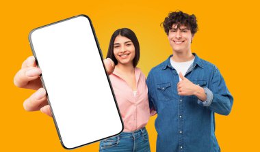 Sarı arka planda genç bir adam ve kadın duruyor. Kadın beyaz ekranlı akıllı telefonu izleyiciye doğru tutarken, erkek de başparmağını kaldırıyor. İkisi de gülümsüyor.