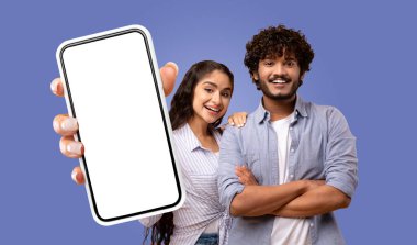 Genç bir Hintli çift, saf mavi bir arka planın önünde duruyor. Kadın kameraya doğru beyaz ekranlı akıllı bir telefon tutuyor. Eli çerçevede görünüyor..