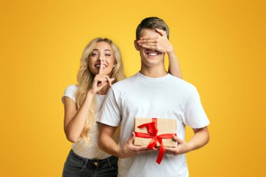 Sürpriz. Kız erkek arkadaşlarının gözlerini kaplayan ve sarı stüdyo arka plan üzerinde sessizlik işareti gösteren