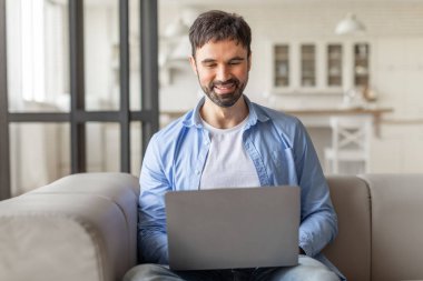 Modern bir oturma odasındaki kanepeye oturup dizüstü bilgisayar kullanan bir adam. O, mavi bir gömlek giyiyor ve ekrana bakarken gülümsüyor. Arka planda cam kapılar var..