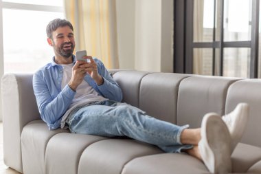 Mavi gömlekli ve kot pantolonlu bir adam gri bir kanepede oturuyor. Rahatlıyor, telefonuna bakıyor ve gülümsüyor. Ayakları kanepede ve beyaz spor ayakkabı giyiyor..