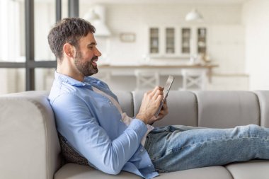 Mavi tişörtlü ve kot pantolonlu bir adam gri bir koltukta rahatça oturur ve tabletin içinde süzülür. Arkasına yaslanıyor ve gülümsüyor, rahat ve mutlu görünüyor. Oda aydınlık ve modern.