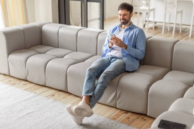 Oturma odasındaki açık gri modüler koltukta oturan bir adam telefonunu kullanırken rahatlamış ve gülümsüyor. Adam mavi düğmeli gömlek, beyaz tişört, kot pantolon ve beyaz spor ayakkabı giyiyor.