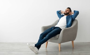 Sakin genç Arap adam, beyaz stüdyo duvarına yaslanmış koltukta dinleniyor, kopyalama alanı. Yakışıklı Orta Doğulu adam huzurlu bir hafta sonu sabahının keyfini çıkarıyor, elleri başının arkasında dinleniyor.