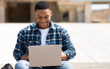 İnternette sörf yapmak. Ön manzara portresi, dizüstü bilgisayarda yazan gülümseyen siyahi kişi, dışarıda parkta oturuyor.