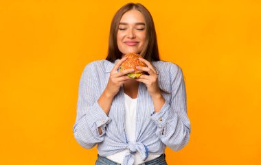 Abur cubur. Sarı Arkaplanda Duran Neşeli Kadın Burger Kokuyor. Stüdyo Çekimi
