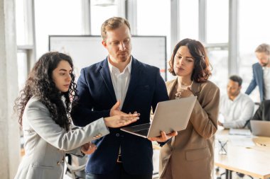 Modern bir ofiste dizüstü bilgisayarın etrafında toplanmış üç iş adamı duruyor. Hepsi dikkatle dinliyor ve üretken bir konuşma yapıyor gibi görünüyorlar..