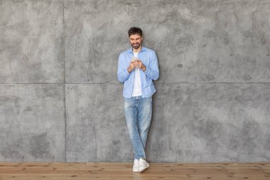 Mavi gömlekli ve kot pantolonlu genç bir adam sırtı beton bir duvara dönük duruyor. Akıllı telefonunu kullanıyor ve ekrandan aşağı bakarken gülümsüyor. Zemin ahşaptan yapılmıştır..