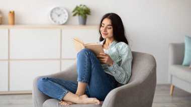 Evde oyalanalım. Milenyum Hindistanlı kadın rahat koltukta oturuyor açık bir kitapla. Güzel genç bayan heyecanlı hikayeler okuyor, tembelliğin tadını çıkarıyor, rahatlatıcı bir hafta sonu geçiriyor.