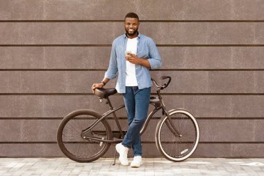 Artık trafik sıkışıklığı yok. Bisikletli yakışıklı Afro-Amerikalı adam gri bina arka planında akıllı telefon kullanıyor.