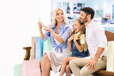 Üç kişilik bir aile alışveriş merkezindeki bir bankta oturuyor, etrafı alışveriş torbalarıyla çevrili. Anne elinde akıllı bir telefonla selfie çekerken baba ve kızı gülümsüyor.