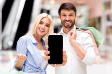 Bir adam ve bir kadın, her ikisi de gülümsüyor, alışveriş merkezinde çantalarla dikiliyorlar, elinde siyah boş ekranlı akıllı bir telefon tutuyorlar.