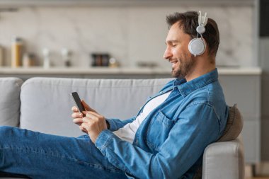 Kulaklık takan bir adam gri bir koltukta oturuyor, akıllı telefonuna bakarken gülümsüyor. Kot gömleğinin altında beyaz bir tişörtü var ve bacakları önünde uzanıyor..