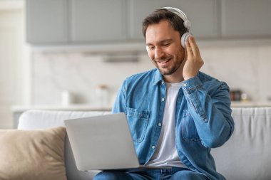 Kulaklık takan bir adam oturma odasındaki koltukta oturup gülümserken ve dizüstü bilgisayar kullanırken. Beyaz bir tişört ve kot pantolonun üzerine mavi bir kot pantolon giyiyor..