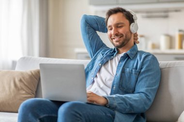 Bir adam açık gri bir koltukta oturuyor, bir eli başının arkasında, kulaklık takıyor ve dizüstü bilgisayar kullanırken gülümsüyor..
