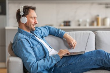 Kot gömlekli bir adam açık renkli bir kanepede oturuyor, kulaklıkları açık ve dizüstü bilgisayarı kucağında. Gülümsüyor, bilgisayarının ekranına bakıyor. Odanın modern bir tasarımı var.