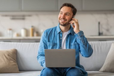 Mavi gömlek ve kot pantolon giyen bir adam oturma odasındaki beyaz bir kanepede oturur. O, kucağında bir dizüstü bilgisayarla gülümsüyor ve telefonla konuşuyor. Kanepede bir yastık var..