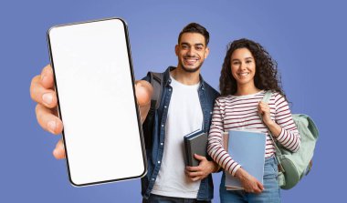 İki Arap öğrenci mavi bir arka planın önünde duruyor. İkisi de gülümsüyor, kitap ve sırt çantaları tutuyor. Adam elinde bir akıllı telefon, önünde de boş bir ekran var..