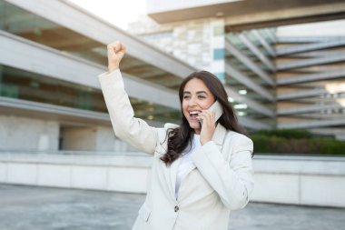 Genç bir iş kadını güneşli bir günde modern bir ofis binasının önünde duruyor. Telefonla konuşuyor. Sağ eli havada. Kutlama jesti yapıyor.