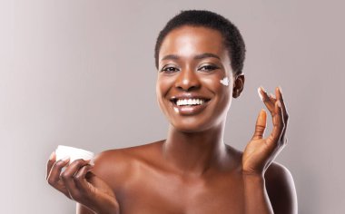 Deri hidrasyonu. Yüze Nemlendirici Krem Tutan ve Uygulayan Neşeli Siyah Kız Portresi, Boş Alanı Olan Gri Arkaplan