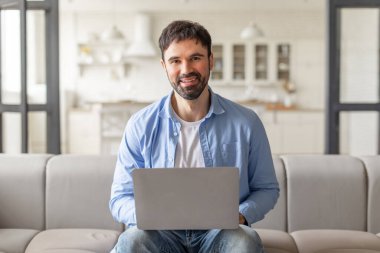 Modern bir evde bir adam bir kanepede oturuyor, dizüstü bilgisayar kullanıyor. Gülümsüyor ve doğrudan kameraya bakıyor. Arka plan bulanık ve açık renkli bir oturma odasını gösteriyor.