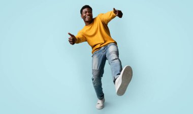 Kaygısız Afro-Amerikan genç adam gündelik kıyafetiyle başparmak kaldırıyor ve bacağını kaldırıyor, mavi stüdyo arka planında poz veren mutlu siyah adam, tam boy çekim, fotokopi alanı olan panorama