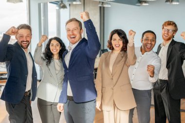 Bir grup iş adamı modern bir ofiste ayakta duruyor, kutlama için hepsi gülümsüyor ve yumruklarını kaldırıyorlar. Başarılı bir projeyi ya da başarıyı kutluyor gibi görünüyorlar..