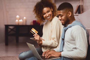 Gülümseyen siyah çift akşam evde kredi kartı ve laptopuyla alışveriş yapıyor.