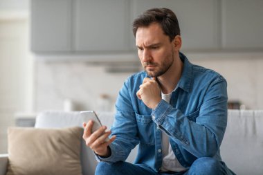 Oturma odasındaki koltukta oturan bir adam akıllı telefonunu okurken endişeli görünüyor. O mavi bir gömlek ve kot pantolon giyiyor. Eli çenesinde ve kaşları çatık..