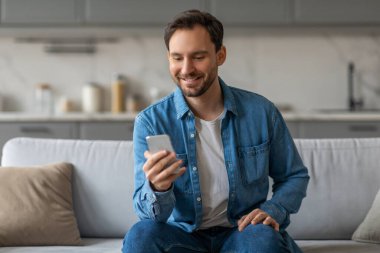 Mavi kot gömlekli bir adam beyaz bir kanepede oturuyor, akıllı telefonuna bakarken gülümsüyor. Kanepe mutfak tezgahının önünde beyaz dolaplarla duruyor..