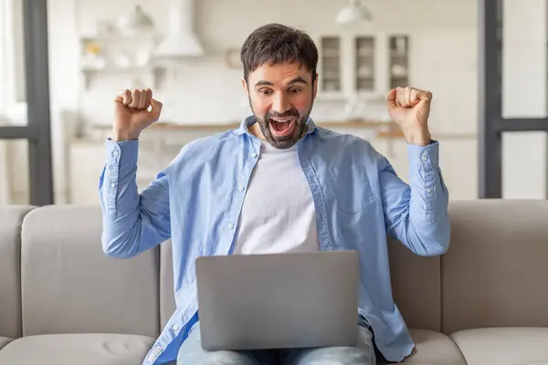 stock image A man sits on a couch, smiling widely and raising both fists in the air, while looking at a laptop screen. He appears to be celebrating a victory or success.